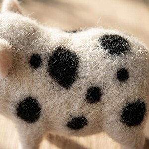 May include: A needle-felted animal figurine crafted from off-white wool fibers, featuring a textured, fuzzy surface with distinct, irregular black spots scattered across its back and side, showcasing the intricate, hand-crafted fiber construction.