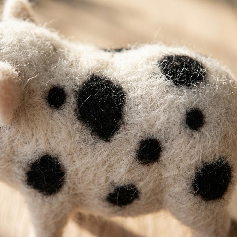 May include: A needle-felted animal figurine crafted from off-white wool fibers, featuring a textured, fuzzy surface with distinct, irregular black spots scattered across its back and side, showcasing the intricate, hand-crafted fiber construction.