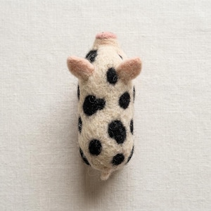 May include: A needle-felted pig figurine crafted from off-white wool, featuring distinct black spots scattered across its body. The small animal figure has two pointed, light pink ears and a rounded, light pink snout at the top end.