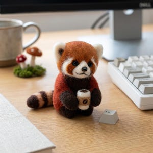 May include: A handcrafted red panda figurine with a coffee cup. The red panda has a brown and white face, red fur, and a striped tail. The figurine is holding a small coffee cup with a coffee bean design.