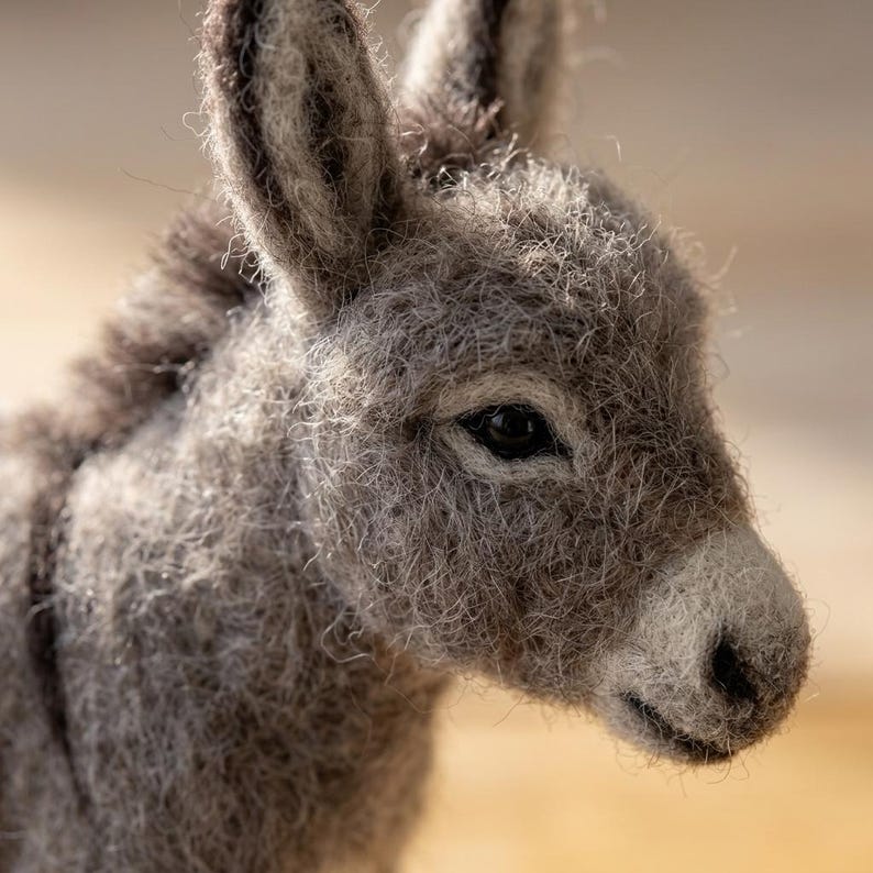 May include: A needle-felted donkey figurine crafted from textured gray wool fibers. The sculpture features upright ears, a dark mane, and a lighter gray muzzle with a black nose and dark, glossy eyes, capturing the animal's head and neck.