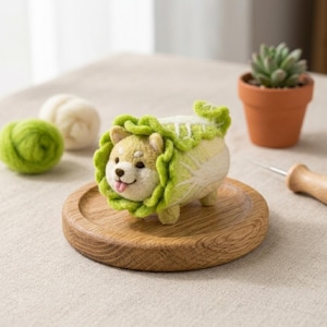 May include: A whimsical felted Shiba Inu dog figurine, crafted with a cabbage-like body and a green leafy collar. The dog has a tan and white face with a pink tongue sticking out. It rests on a small wooden coaster, with a succulent in a terracotta pot in the background.