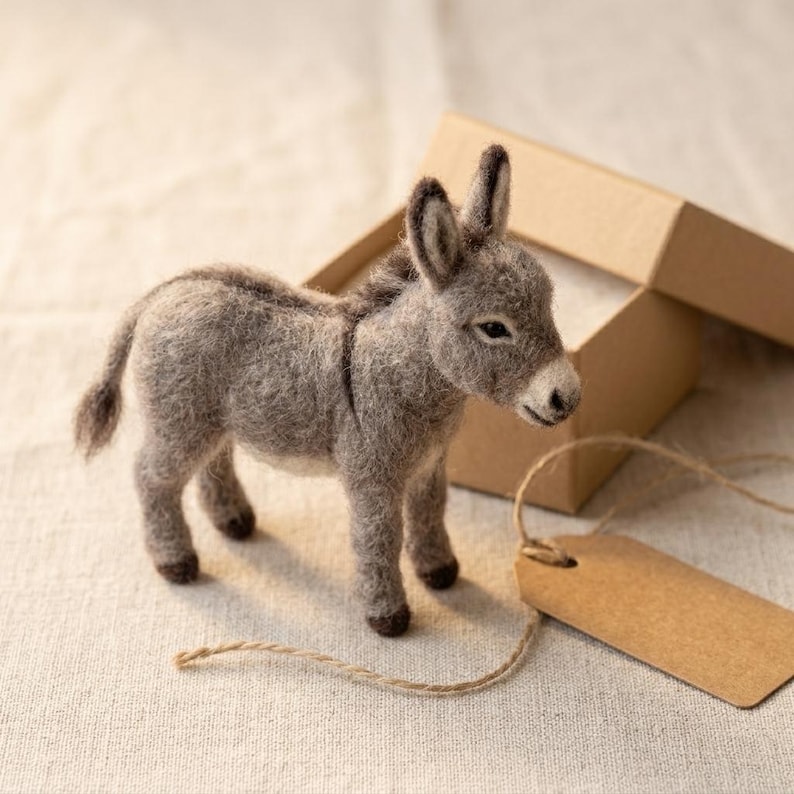 May include: A needle-felted gray donkey figurine with a textured wool finish, upright ears, and a small tail. The donkey stands in front of an open brown cardboard gift box and a blank brown paper tag attached by a thin string.