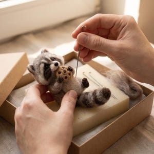 May include: A needle-felted raccoon figurine made of grey, white, and black wool fibers. The raccoon has a striped tail and is holding a small, round, tan-colored cookie with dark brown spots. A metal felting needle is positioned near the figure.