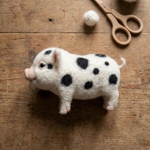 May include: A needle-felted pig figurine made of off-white wool with distinct black spots across its body, a pink snout, and a small curled tail. The pig is positioned near a small ball of white wool and a pair of light-colored wooden scissors.