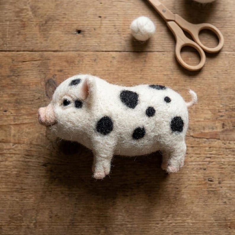 May include: A needle-felted pig figurine made of off-white wool with distinct black spots across its body, a pink snout, and a small curled tail. The pig is positioned near a small ball of white wool and a pair of light-colored wooden scissors.