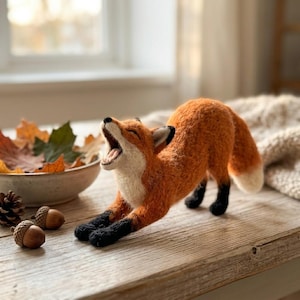 Op de afbeelding: Handgemaakte oranje vossenfiguur met zwarte poten en oren, gapend. De vos rekt zich uit op een houten oppervlak, met een kom herfstbladeren en eikels in de buurt. De vos heeft een witte borst en staartpunt.