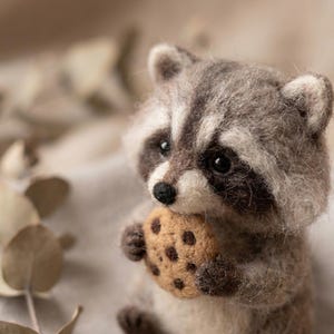 May include: A needle-felted raccoon figurine crafted from fuzzy gray and white wool fibers. The raccoon has dark eye patches, a small black nose, and pointed ears. It is holding a round, tan-colored felted cookie with dark brown chocolate chips.