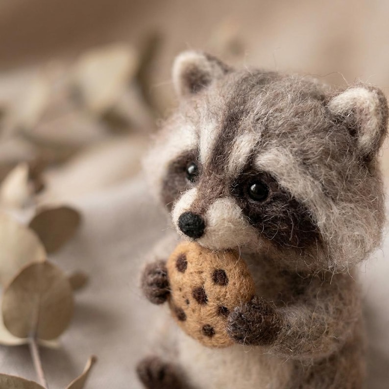 May include: A needle-felted raccoon figurine crafted from fuzzy gray and white wool fibers. The raccoon has dark eye patches, a small black nose, and pointed ears. It is holding a round, tan-colored felted cookie with dark brown chocolate chips.