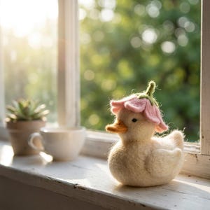 May include: A needle-felted cream-coloured duck figurine wearing a pink flower-shaped hat with a green stem on top. The duck has a small orange beak and dark eyes, crafted from soft wool fibres with a textured appearance and rounded shape.