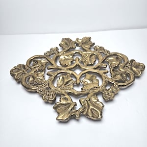 May include: A gold-toned metal trivet featuring an intricate openwork design of embossed grape leaves and small flowers. The decorative piece has a symmetrical, elongated diamond shape with a polished finish and a detailed, textured surface.