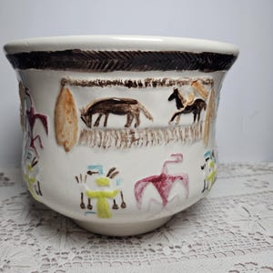 May include: A white ceramic planter featuring a dark brown textured rim and hand-painted illustrations of horses, figures in yellow and red attire, and abstract shapes. The bowl-shaped vessel has a smooth finish and a wide, flared opening.