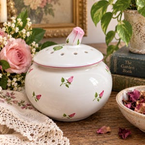May include: A white ceramic potpourri jar with a rounded body and a removable lid featuring a sculpted pink rosebud handle. The jar is decorated with small painted pink rosebuds and green leaves, and the lid has several circular ventilation holes.