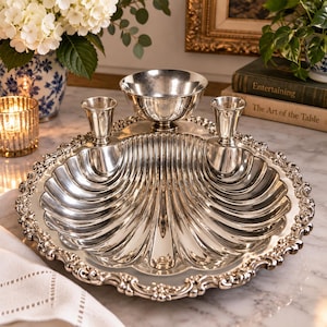 May include: A silver-plated serving tray shaped like a large scallop shell with a decorative scalloped edge. The tray features two attached cylindrical cups and a central pedestal bowl, all finished in a polished, reflective silver tone.
