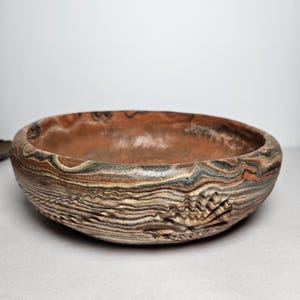 May include: A shallow, round ceramic bowl featuring a marbled, multi-colored pattern of brown, black, tan, and cream swirls. The exterior surface displays a textured, relief-carved shell motif integrated into the layered, wavy design.