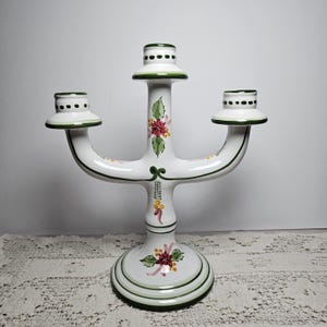 May include: A white ceramic candelabra with three candle holders, featuring hand-painted green trim and floral motifs including red poinsettias and green holly leaves. The base is circular with green rings, and the candle cups have dotted green rims.