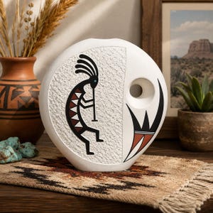 May include: A round white ceramic vase featuring a textured surface and a black, white, and terracotta Kokopelli figure playing a flute. The vase includes a circular cutout on the right side and a stylized black and terracotta geometric design.