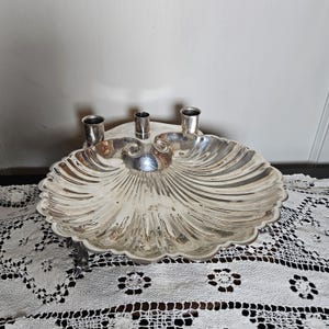 May include: A silver-toned metal dish shaped like a large scallop shell, featuring three small cylindrical candle holders attached to the back edge. The dish has a scalloped rim and a ribbed surface texture, supported by small decorative metal feet.
