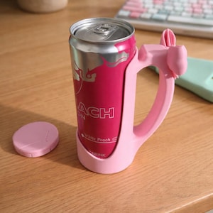 以下が含まれることがあります： 銀色の蓋が付いたピンク色のドリンク缶が、お揃いのピンク色のハンドルで保持されています。「ACH」と「White Peach Drink」の文字が印刷されています。左側にはピンク色のコースターがあります。
