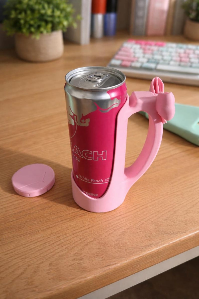 May include: A pink drink can with a silver top is held in a matching pink handle. The can has the word "ACH" and "White Peach Drink" printed on it. A pink coaster sits to the left.
