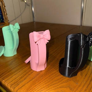 May include: Three decorative phone holders in pastel colors: mint green, pink, and black. Each holder features a bow accent at the top. The holders are displayed on a wooden surface.