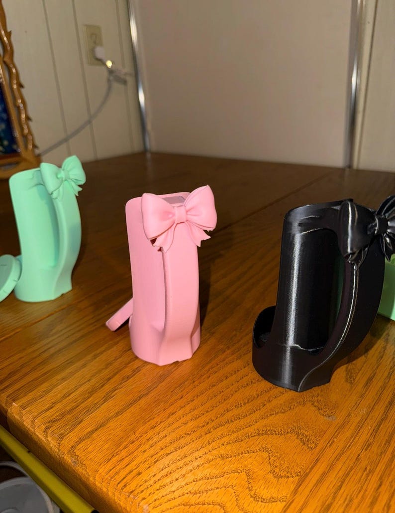 May include: Three decorative phone holders in pastel colors: mint green, pink, and black. Each holder features a bow accent at the top. The holders are displayed on a wooden surface.