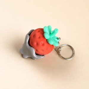 Pode incluir: Um porta-chaves de morango vermelho com caule e folhas verdes. O porta-chaves está preso a um suporte prateado e a um anel de metal. O morango tem uma superfície texturizada.
