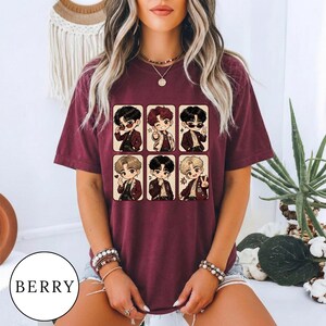 Op de afbeelding: Een bessenkleurig T-shirt met korte mouwen en een grafische print van zes verschillende personages in chibi-stijl, gerangschikt in een raster. Elk personage is afgebeeld in een andere pose en draagt een bruin jasje over een wit overhemd.