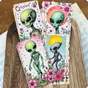 Peut inclure: Quatre cartes rectangulaires présentant des illustrations colorées d'êtres extraterrestres. Chaque carte affiche un personnage alien unique entouré de fleurs roses, d'étoiles et de fonds célestes. Une carte inclut le texte Queen en haut.