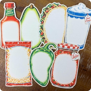 May include: A collection of six die-cut paper notepads shaped like food items, including a hot sauce bottle, an avocado, a taco, a sour cream container, a bag of shredded cheese, a jalapeño pepper, and a jar of salsa with labels.