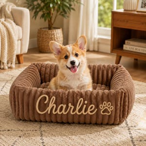 May include: A rectangular brown pet bed with a ribbed texture, featuring the name Charlie embroidered in a light tan script font alongside a paw print icon. A tan and white Corgi puppy sits inside the soft, plush, high-walled pet bed.