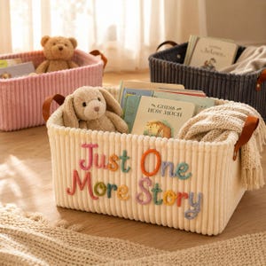 Könnte beinhalten: Ein cremefarbener Aufbewahrungskorb aus Kord mit braunen Ledergriffen, verziert mit dem gestickten Text "Just One More Story" aus buntem Garn. Der Korb enthält ein Plüsch-Hasenspielzeug, Kinderbücher und eine strukturierte Strickdecke.