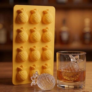 May include: A yellow silicone pineapple-shaped ice cube tray with 12 molds. A clear pineapple ice cube is in a glass of amber liquid. A clear, spherical ice ball and a glass of amber liquid are also present.