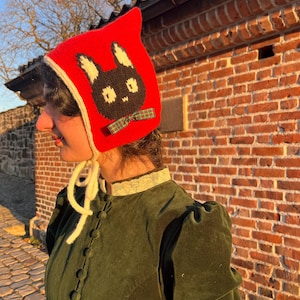 Op de afbeelding: Een rode gebreide muts met een zwart konijnengezicht en een groene geruite strik. De muts heeft witte randen en linten. De persoon op de foto draagt een donkergroene fluwelen jas met een kanten kraag.