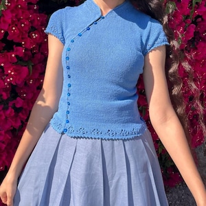 May include: A short-sleeved blue knit top featuring a mandarin collar and an asymmetrical front closure secured with a vertical row of small blue buttons. The garment has a decorative openwork lace pattern along the hem and sleeve edges.
