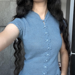 May include: A dusty blue knit top featuring a mandarin collar, short sleeves with scalloped edges, and a vertical front closure with six round buttons secured by decorative, flower-shaped loops. The hem also features a delicate lace pattern.