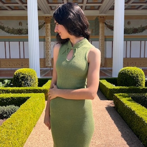 Peut inclure: Robe sans manches vert olive avec une encolure en trou de serrure. La robe présente un motif en tricot texturé et un ourlet au genou. La robe est portée à l'extérieur dans un jardin avec des haies taillées et une architecture classique en arrière-plan.