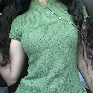 May include: A sage green knit top featuring a mandarin collar, short sleeves, and an asymmetrical button-down placket along the upper left chest. The garment includes a delicate openwork lace pattern along the sleeve cuffs and the bottom hemline.