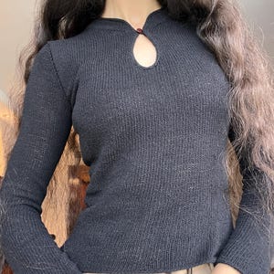 May include: A black long-sleeved knit jumper featuring a ribbed texture, a high neckline, and a small keyhole cutout at the centre of the chest secured by a single dark red button. The garment has a fitted silhouette and long sleeves.