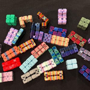 May include: A collection of colorful fidget toys in various shapes and color combinations. The toys are made of small, connected blocks in shades of green, blue, pink, orange, and purple. The background is a dark, solid color.