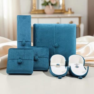 Puede incluir: Una colección de joyeros de terciopelo color turquesa en varias formas y tamaños. Las cajas se cierran con un botón. Dos joyeros abiertos muestran anillos con piedras preciosas. El fondo incluye una superficie blanca y un espejo decorativo borroso.