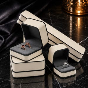 May include: A collection of cream-colored jewelry boxes with black trim and dark gray velvet interiors. One square box holds two rings, while a smaller square box holds one ring. A long, rectangular box is positioned behind the square boxes.