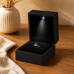May include: A matte black square ring box with a hinged lid and a soft black velvet interior. The box features a small integrated LED light at the top that illuminates a silver ring with a single blue gemstone centered in the velvet slot.