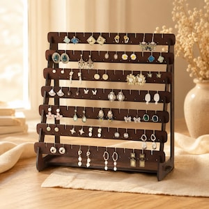 Peut inclure: Un présentoir à boucles d'oreilles en bois brun foncé avec plusieurs rangées horizontales. Le présentoir est rempli de diverses boucles d'oreilles de différents styles et couleurs, notamment des clous, des créoles et des boucles d'oreilles pendantes. Les boucles d'oreilles sont exposées sur une surface claire.