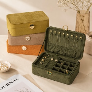May include: A stack of three rectangular jewelry boxes in olive green, taupe, and orange, with gold-tone hardware. The open, dark green velvet jewelry box displays rings and compartments for jewelry storage. A small dish of pearls and a magazine are in the foreground.
