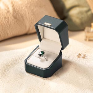 Puede incluir: Una caja de anillo verde oscuro con un anillo plateado con una piedra preciosa verde oscuro. La caja está abierta, revelando un interior blanco. La caja está sobre una superficie color crema, con un fondo borroso.