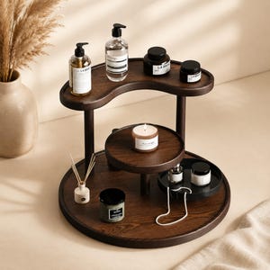 Peut inclure: Un présentoir à étages en bois brun foncé avec une étagère supérieure en forme de croissant et une base circulaire. Il contient divers flacons de soins, des petits pots, une bougie parfumée, un diffuseur à bâtonnets et un collier de perles blanc.