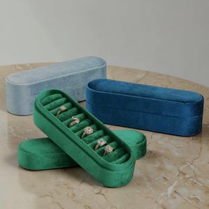 May include: Velvet ring boxes in blue and green. The open green box displays rings. The oblong boxes have a rounded top and a velvety texture. The boxes are on a marble surface. The boxes are approximately 20 cm long.
