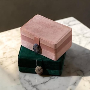 Pode incluir: Duas caixas de joias de veludo, uma empilhada em cima da outra. A caixa superior é rosa empoeirado com uma faixa rosa mais escura e um fecho de pedra. A caixa inferior é verde escuro com um fecho de pedra. Ambas as caixas são retangulares.