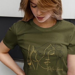 May include: Olive green t-shirt featuring a gold line art design of a woman's face, a wine glass, and the text "Spita-fina". The design is centered on the front of the shirt. The model is wearing dark wash jeans.